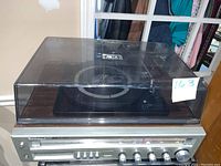 Front view of the turntable unit with clear plastic dust cover closed, showing the faux wood finish and control panel with knobs and buttons, condition visible including scratches and minor damage.