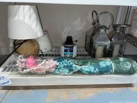 Photo showing the arrangement of a small lamp with a small fabric lampshade and coiled rope base, three silver lanterns with candles inside, tiki light, grass Hawaiian skirt wrapped in plastic, additional small lampshade, and some lamp oil or fuel container.