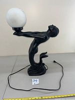 Full side view of black resin mermaid lamp holding white globe light. Power cord attached and lying on surface. Tail detached and in need of repair.