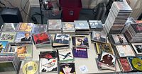 Photo showing multiple stacks and loose CDs arranged on a table, featuring a variety of album covers from different music genres and artists, including some loose discs and handwritten CDs visible.