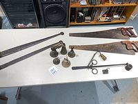 Top view photo showing two long iron strap hinges, five small bells, two wooden handled saws, and a set of weights laid out on a table.