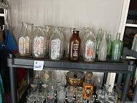 Front view of shelf showing milk bottles with various orange and black labels plus green glass and brown Arrow Lakes Dairy bottles