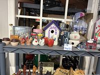 Shelf display of vintage home decor items including bird house oil lamp, potpourri pot, collectible wooden house, RBG action figure, drinking cup, glass canister, glass fish, galvanized tin planter, salt and pepper shakers.