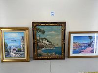 Three framed artworks against white wall: left is San Francisco trolley painting, center Mediterranean coastal scene painting, right Padova villa print