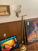 Full side view of the antique brass floor lamp with milk glass shade standing next to various framed art pieces.