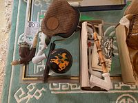 Top-view photo showing multiple antique pipes, a decorative tobacco box with horse and rider embossed design, a brown textured women's clutch bag, and some pipe accessories on a patterned rug.