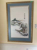 Framed Asian needlework art on cloth showing mountains, trees, fisherman, and calligraphy in muted colors with gold decorative frame.