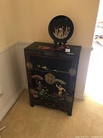 Black Asian-style painted cabinet with painted floral and figure scenes, metal hinges and latches, standing in a corner with a decorative round Japanese tray on a wooden stand on top.
