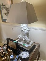 Front view of the table lamp showing the white ceramic Foo Dog base and beige cloth shade, placed on a wooden table among other decorative items.