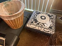 Photo showing both wicker wastebasket with plastic liner and blue upholstered footstool with white pattern and wooden legs placed on brown carpet.