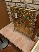 Angled view of the brass fireplace screen showing the embossed ship design and decorative border. The handle and legs are visible.