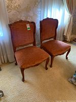 Pair of antique chairs with rust-orange padded upholstery, wide carved wooden frames, and cabriole legs placed side by side on beige carpeted floor near window with sheers and floral wallpaper.