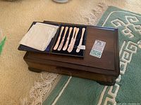 Closed box with silver-plated butter spreaders inside and a folded white cloth, displayed on a wooden table and patterned carpet.