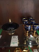 Photo of six cobalt blue and gold decorative Judaica goblets displayed upright with intricate white designs and Jewish motifs including a Star of David and Moses figure, next to a covered bowl with lid, all on glass shelf.