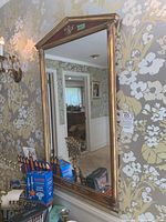 Front view of the tall rectangular mirror with rustic gold frame and reddish triangular pediment top, hanging on floral wallpapered wall.