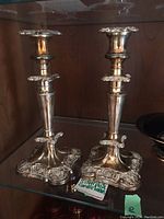 Pair of silver-plated candle holders on glass shelf showing ornate bases and platforms for candles