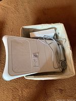 White Nintendo Wii console model RVL-001 laying sideways in a soft beige container, includes cables and power adapter.