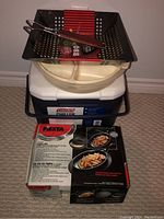 Photo showing stacked items including Coleman cooler at bottom, white divided pan from Fiesta fajita kit in middle, and black metal BBQ grill basket with wooden handled tongs on top.