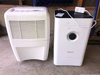 Two air treatment units side by side on a garage floor: a white Whirlpool dehumidifier and a white Boneco air cleaner with black top and dial.