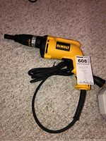 Yellow and black DeWalt corded drywall screwdriver with power cord coiled beside it, lying on carpet. Visible DeWalt label and attached Masco tag.