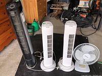 Four fans displayed in garage: one black tower fan, two white tower fans, one white traditional fan.
