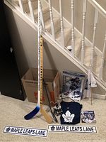 Overview photo showing two hockey sticks, two street signs, a shirt, a flag, a small poster, a figure lamp, and a small hockey figurine placed on carpet near stairs.
