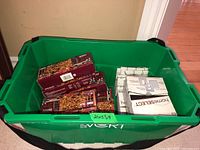 Green plastic storage bin containing three boxes of Emerald indoor clear mini holiday lights and multiple boxes of HomeSelect switch plates.