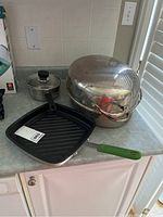 Photo showing the large stainless steel roasting pan with dome-shaped stained lid and rack, next to a square black NordicWare frying pan with green handle, and a small stainless steel pot with glass lid.