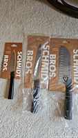 Three Schmidt Bros knives: paring, utility, and Santoku, all in original packaging with cardboard backing and plastic wrap.