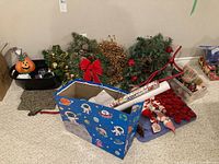 Photo showing multiple Christmas wreaths with decorations, a Halloween pumpkin container, gift wrap rolls in a blue space-themed box, and other seasonal decor items spread on carpet.
