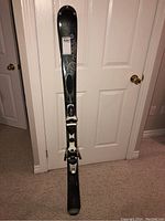 Full length photo of one Dynastar ski standing upright showing the top surface and bindings, against a white door.