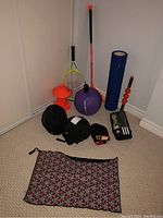 Photo showing sports equipment including yellow and black tennis racket, orange mini pylons, purple twist ball, blue foam roller, orange massage stick, black protective helmets, protective pads, and patterned bag.
