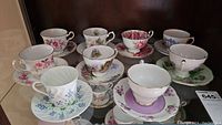 Photo displaying all 9 teacups and saucers set arranged in two rows on glass shelf, showing floral patterns and various colorful designs