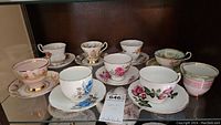 Photo showing front view of 9 assorted teacups and 8 saucers with floral and gold rim patterns on glass shelf