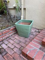Green square pot placed on red brick patio next to a wall.
