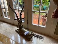 Full side view of the reclining brass/bronze reindeer sculpture placed near a window on tiled floor to show size and shape.