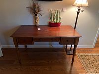 Wooden desk with three drawers, vase with dried plants, red container with white flowers, and floor lamp on the right