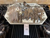 Top-down view of silverware and serving tray on stove, showing various forks, knives, spoons in plastic sleeves and loose, along with large serving ladle and spoons on a rectangular tray.