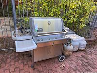 Full view of stainless steel grill with fold-out side shelves, two wheels and three propane tanks