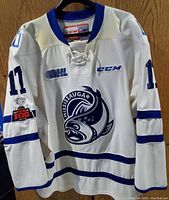 Front view of white hockey jersey with blue stripes, Mississauga Steelheads logo featuring a steelhead fish, and patches on sleeves.