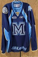 Front view of navy blue and light blue hockey jersey with large M logo and Majors text, team patches on sleeves.
