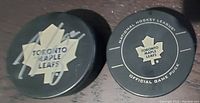 Two black hockey pucks, one with Toronto Maple Leafs logo and official game puck markings, the other with NHL logo and Tyler Bozak #42 label.