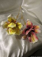 Two large hand painted iris flower sculptures, one yellow and one pink, with detailed petals and gold-colored stems, placed on silky fabric.