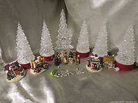 Photo displaying 6 luminary Christmas trees including 5 plastic white trees and 1 central clear glass tree, along with miniature nativity scene figures and 4 small Christmas houses.