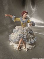 Front angle of antique Dresden porcelain figurine showing seated lady in yellow dress with lace details and crossed legs.
