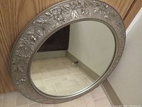 Front view of round mirror showing detailed embossed silver floral and leaf frame surrounding clear reflective glass surface.