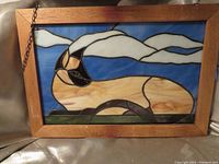 Front view of stained glass sun catcher featuring a Siamese cat lying on grass with a blue sky and clouds background, wooden frame and hanging chain included.
