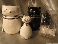 Photo showing the front view of two cat-shaped ceramic storage jars (white and black), a white cat timer, and packaged black and white cat salt and pepper shakers.