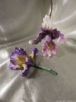 Two large hand painted enameled purple iris flowers shown against a white fabric backdrop. Both flowers feature detailed purple and white petals with gold-tone stamens and green stems.