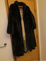 Front and partial side view of the coat hanging on a door, showing its plush dark fabric and open front style with inner lining visible.